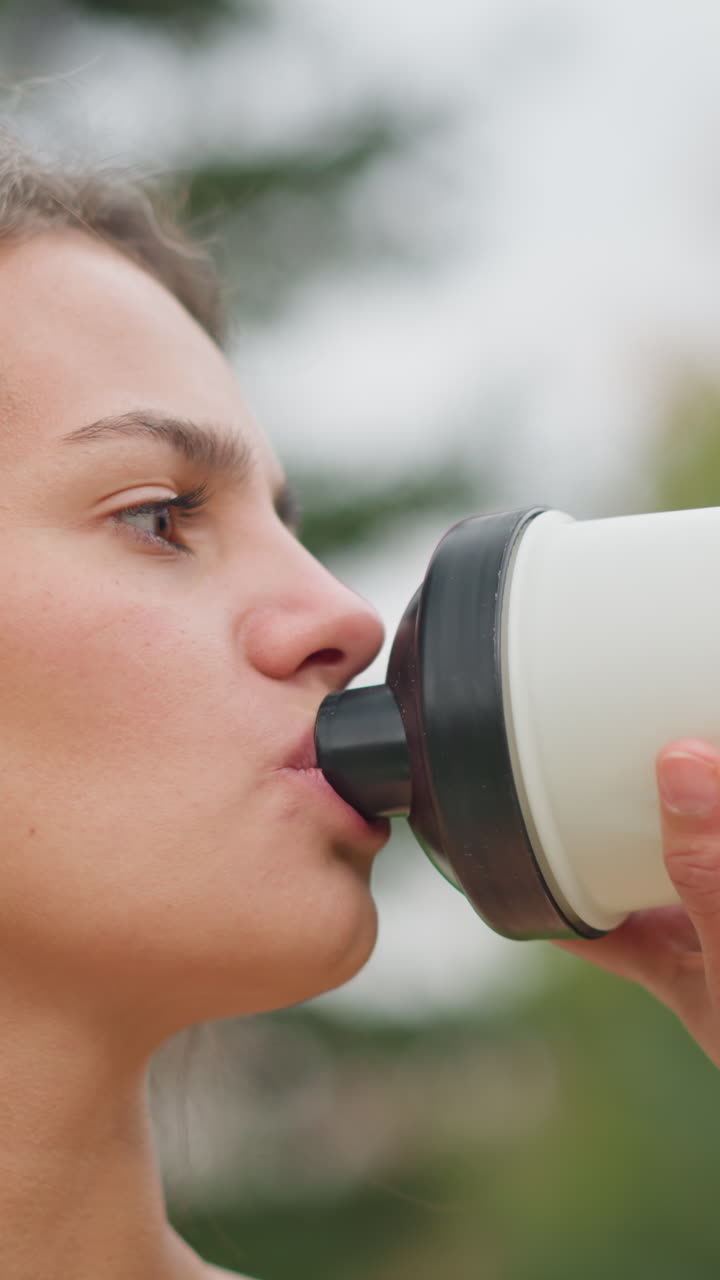 Ritratto di una donna che beve acqua dalla sua bottiglia, concentrandosi sull'idratazione durante l'attività all'aperto; la sua attenzione nel rinfrescarsi sottolinea le routine di benessere e fitness.