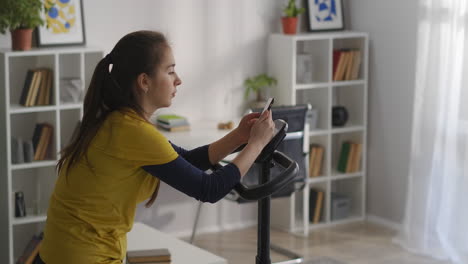 Una-Mujer-Con-Un-Teléfono-Inteligente-Está-Sentada-En-Una-Bicicleta-Estática-En-Casa-Viendo-Redes-Sociales-Y-Eligiendo-Música-Para-Hacer-Ejercicio,-Un-Estilo-De-Vida-Deportivo-Y-Saludable