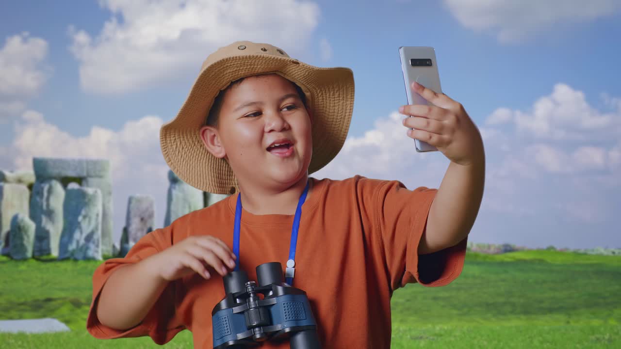 Asian Boy With A Hat And Binoculars Waving Hand Having Video Call On Smartphone While Traveling In Stonehenge. Boy Researcher Examines Something, Travel Adventure, Close Up