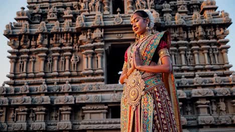 Young woman wearing traditional indian clothing praying with hands together in front of a richly decorated ancient stone temple at sunset, showcasing the rich culture and spirituality of India