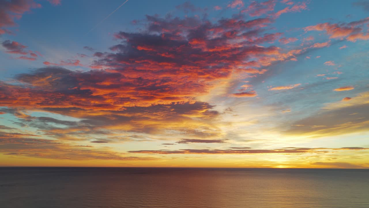 Dronebillede fra luften af en spektakulær solopgang med orange og røde skyer der spejler sig i havet. Plads til tekst.