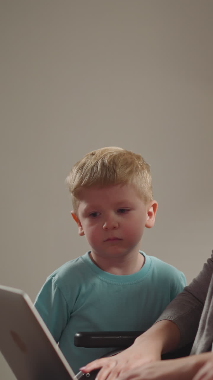 Chubby toddler boy and long-haired preschooler girl watch mommy working on laptop. Freelance woman with muscular weakness sits in wheelchair closeup