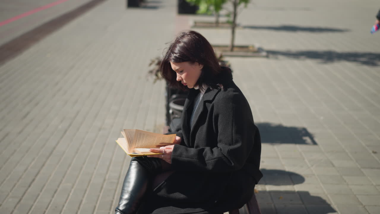 Lady sitting outdoors with crossed legs, reading thoughtfully as wind gently blows and hair flutters, shadow cast on ground, calm, peaceful moment in an urban setting with soft sunlight