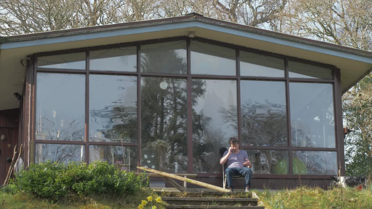 Young man on phone under cabin porch breaks wood in anger