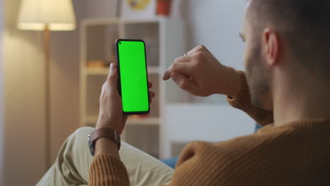 Un-Hombre-Adulto-Está-Viendo-Fotos-En-Un-Teléfono-Inteligente-Deslizando-El-Dedo-En-La-Pantalla-Verde-De-Una-Aplicación-De-Gadget-Y-Un-Sitio-Para-Servicio-Y-Entretenimiento-Descansando-En-Casa-Por-La-Noche