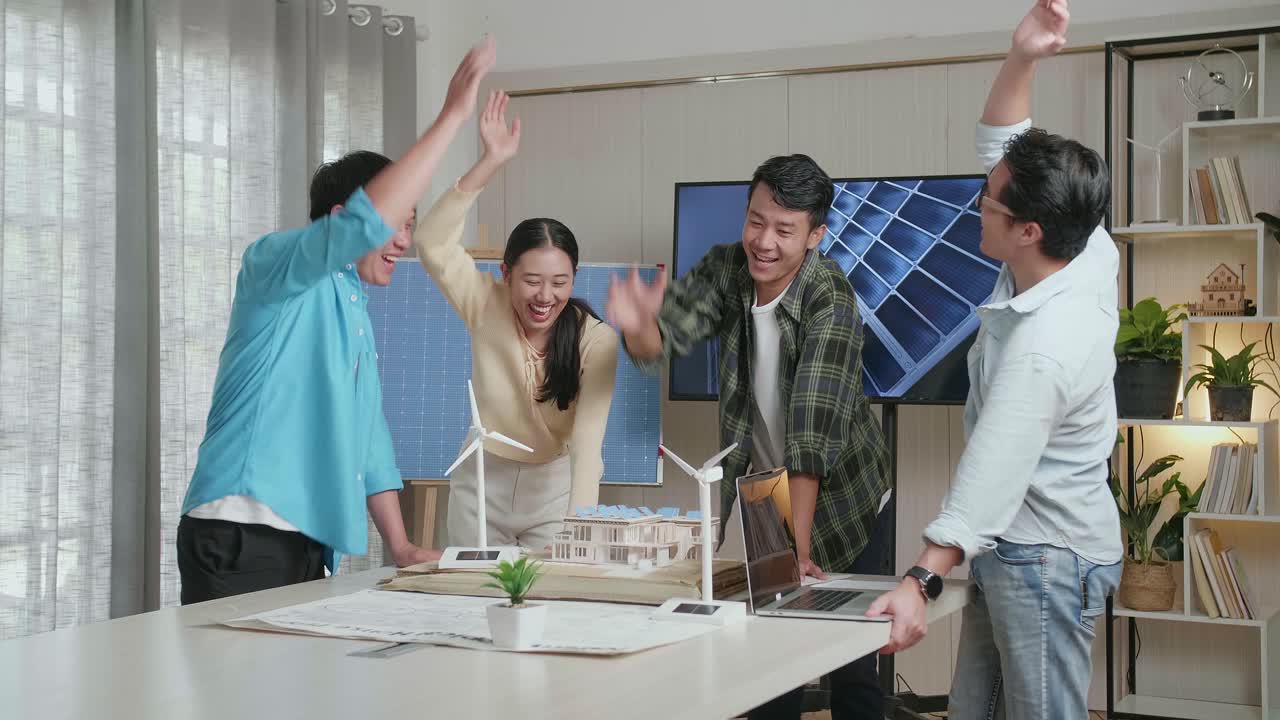 아시아 엔지니어 그룹이 사무실에서 태양 전지판을 가진 작은 집 모델을 작업하는 동안 함께 손을 고 있습니다.