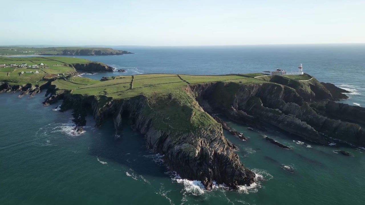 Oprørt hav langs det buede, barske kystområde i West Cork. Luftfoto over Galley Head fyrtårn og klipper med et bredt panorama af marker og landsbyer.