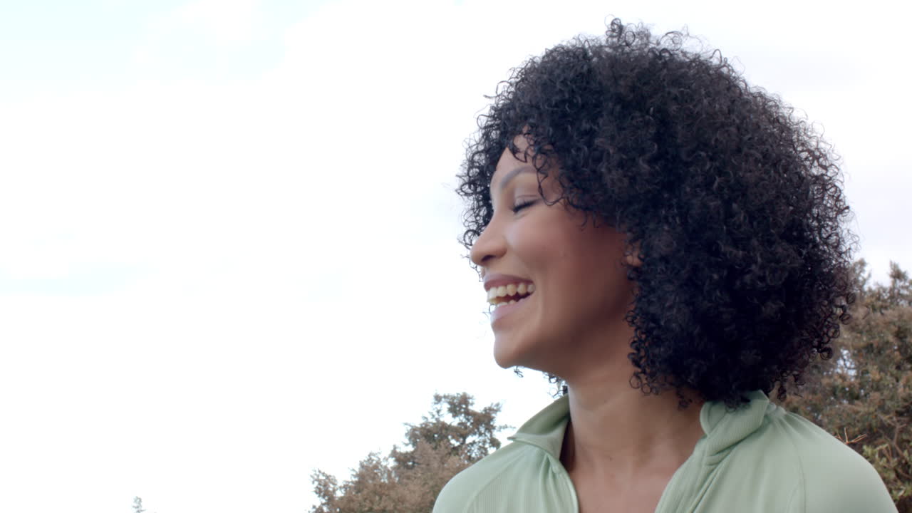 Mulher birracial sorrindo ao ar livre com árvores e céu limpo, com espaço para texto.