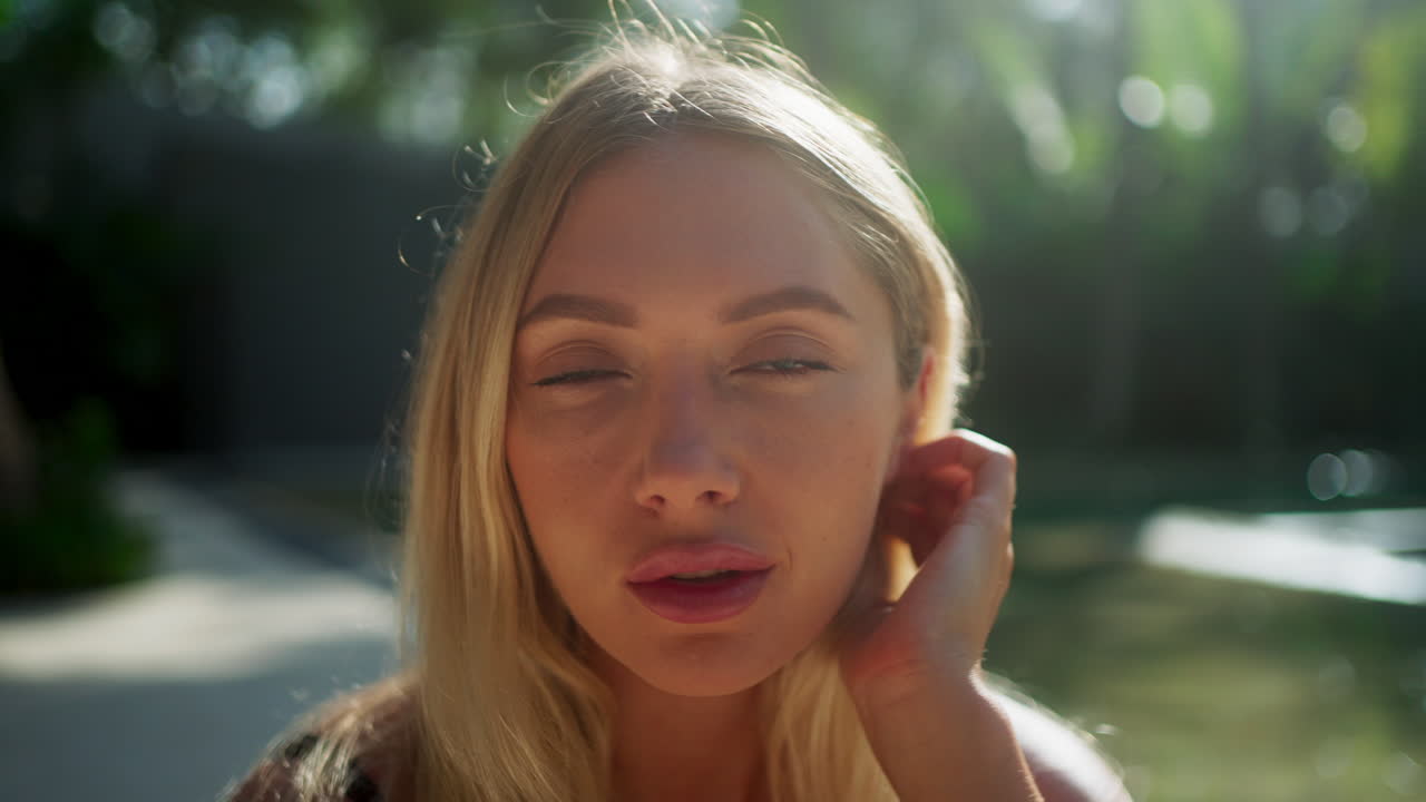 Flirtatious gesture of pretty, blonde, young caucasian model pushing hair behind ear in warm tropical environment next to luxury pool in sunlight - Close up, shallow depth of field, slow motion.