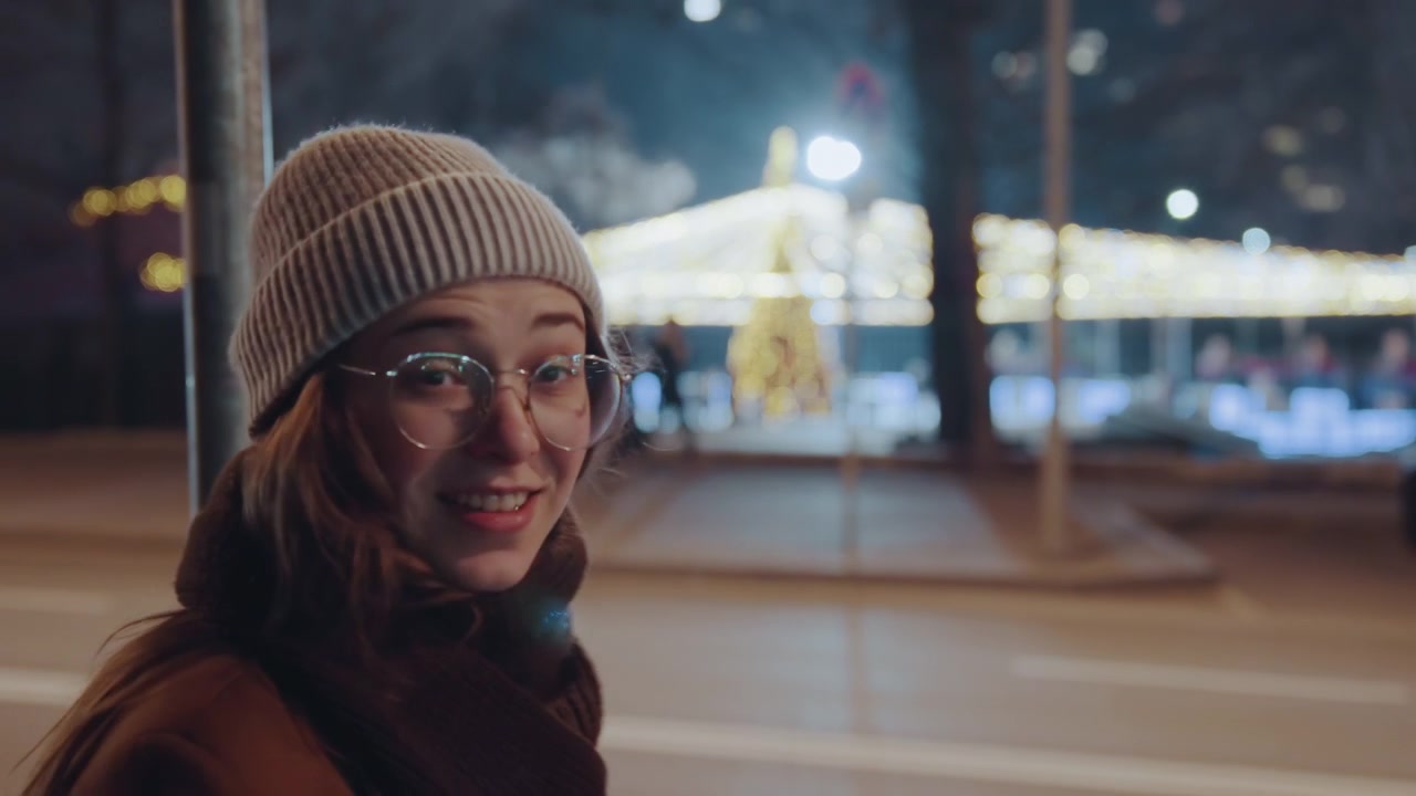 Giovane donna caucasica che si meraviglia delle luci su un viale cittadino, con berretto e occhiali, si copre la bocca e poi indica, energia giocosa da vlogger, bokeh festivo, passeggiata serale fredda, scene di reazioni urbane spontanee.