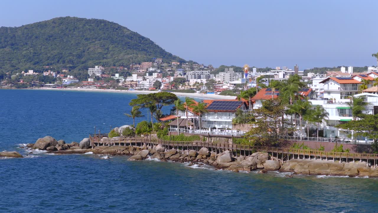 Aerial view capturing Bombinhas in Santa Catarina, Brazil, showcasing a rocky coastline, buildings, a wooden walkway, lush vegetation, and a tranquil blue sea under a clear sky