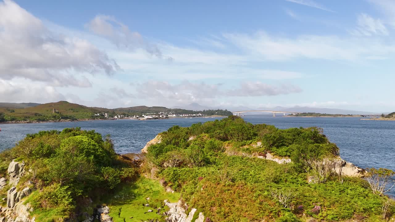Drone glides above rocky green islet in Loch Alsh, revealing scenic coastal landscape in daylight