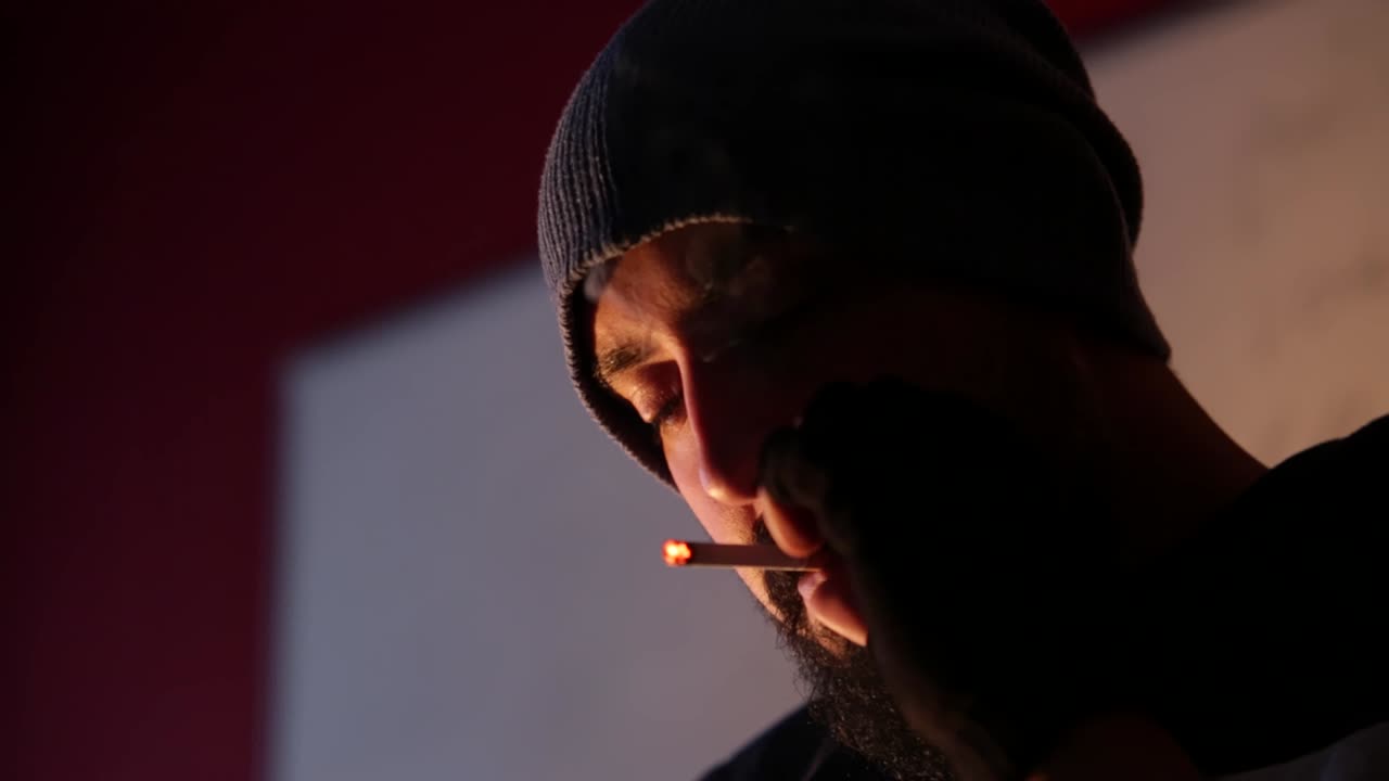 Young bearded man with hat and gloves lighting cigarette and smoking indoor in dark area with blurry background, copy space