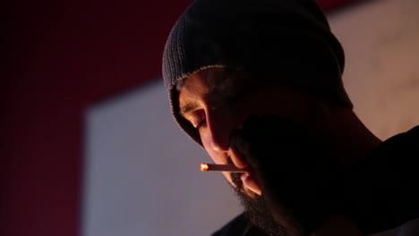 young bearded man with hat and gloves lighting cigarette and smoking indoor in dark area with blurry background, copy space