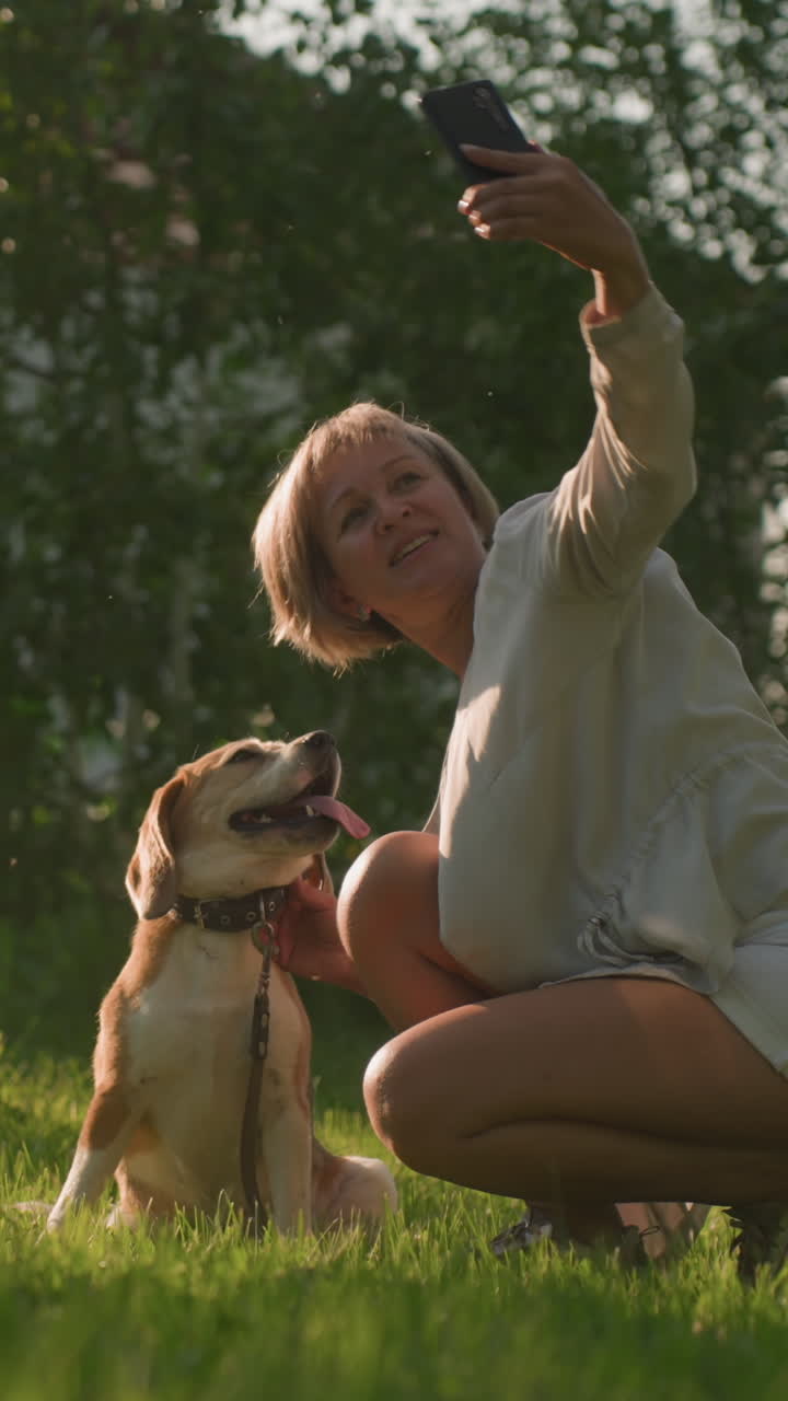 빛 날씨 아래 울창한 초록색 정원에서 즐거운 개와 함께 셀피를 찍는 여성, 건물과 나무가 배경으로, 개는 주인이 에 누워있는 동안 행복하게 숨을 쉬고 앉아 있습니다.