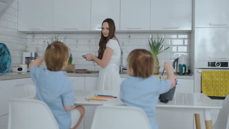 Mom-in-the-kitchen-washing-dishes-and-two-sons-sitting-at-a-Desk-drawing-on-paper-with-colored-pencils.-Young-family-mom-smiles-and-looks-at-children