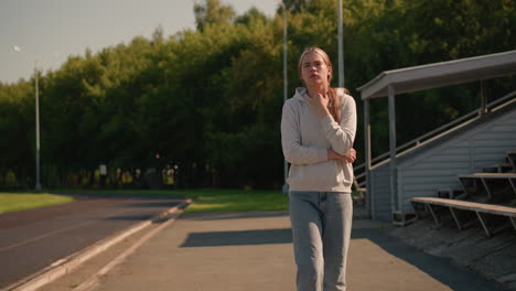 young lady in casual hoodie walks thoughtfully near empty stadium with hand on her hoodie, surrounded by lush green trees and electric poles