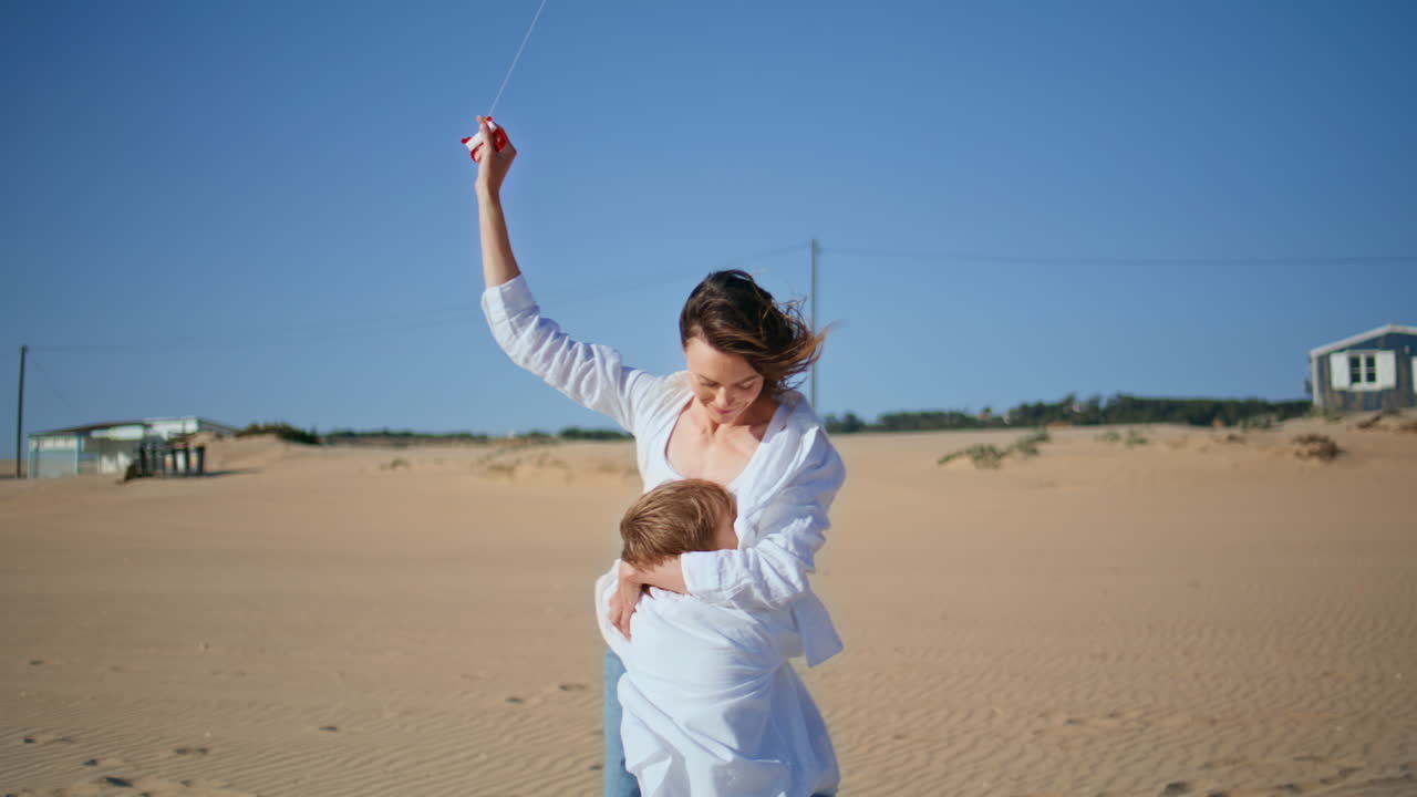 Playful mother child hugging flying kite on sunny beach. Smiling mom holding toy