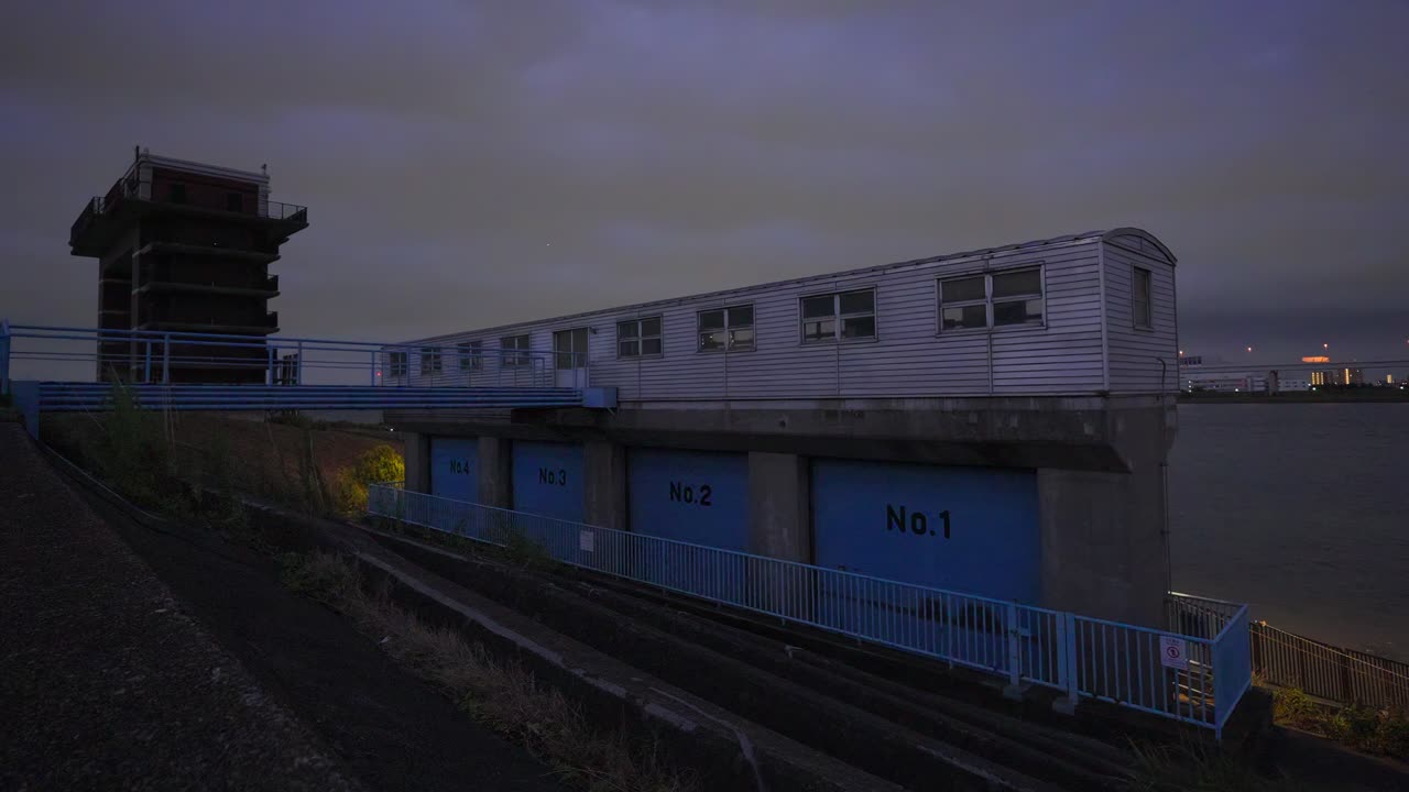 An eye-level shot of the Arakawa Lock Gate's control building at night, with the river and city lights visible in the distance under a starry sky