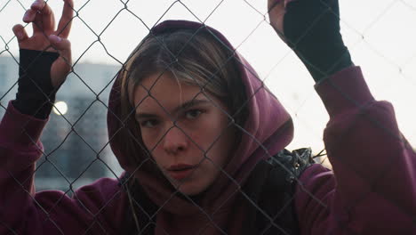 wellness advocate in maroon hoodie leans against chain link fence with hands gripping wire, eyes closed in restful pause after intense outdoor workout at dusk against urban skyline backdrop