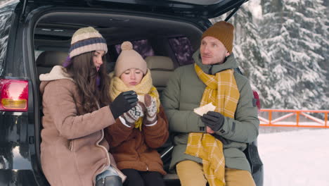 parents and daughter eating sandwiches and drinking hot drink sitting in the trunk of the car in a snowy forest 1