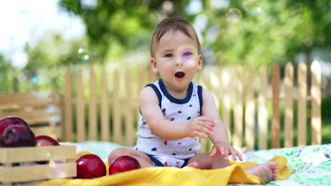 cute baby sitting in the summer garden looks at soap bubbles flying by. toddler sits still and then wants to reach the camera. blurred backdrop.