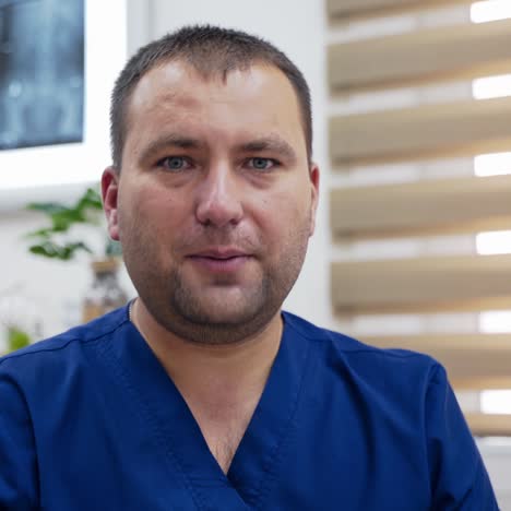 portrait of a male therapist. skilled doctor in blue medical uniform talking on camera in modern medical office. medical specialist consults patients through video call.