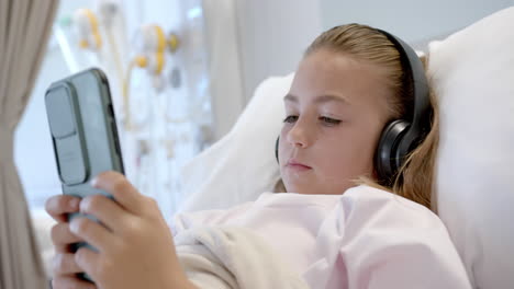 una paciente caucásica acostada en una cama de hospital con auriculares usando un teléfono inteligente, cámara lenta
