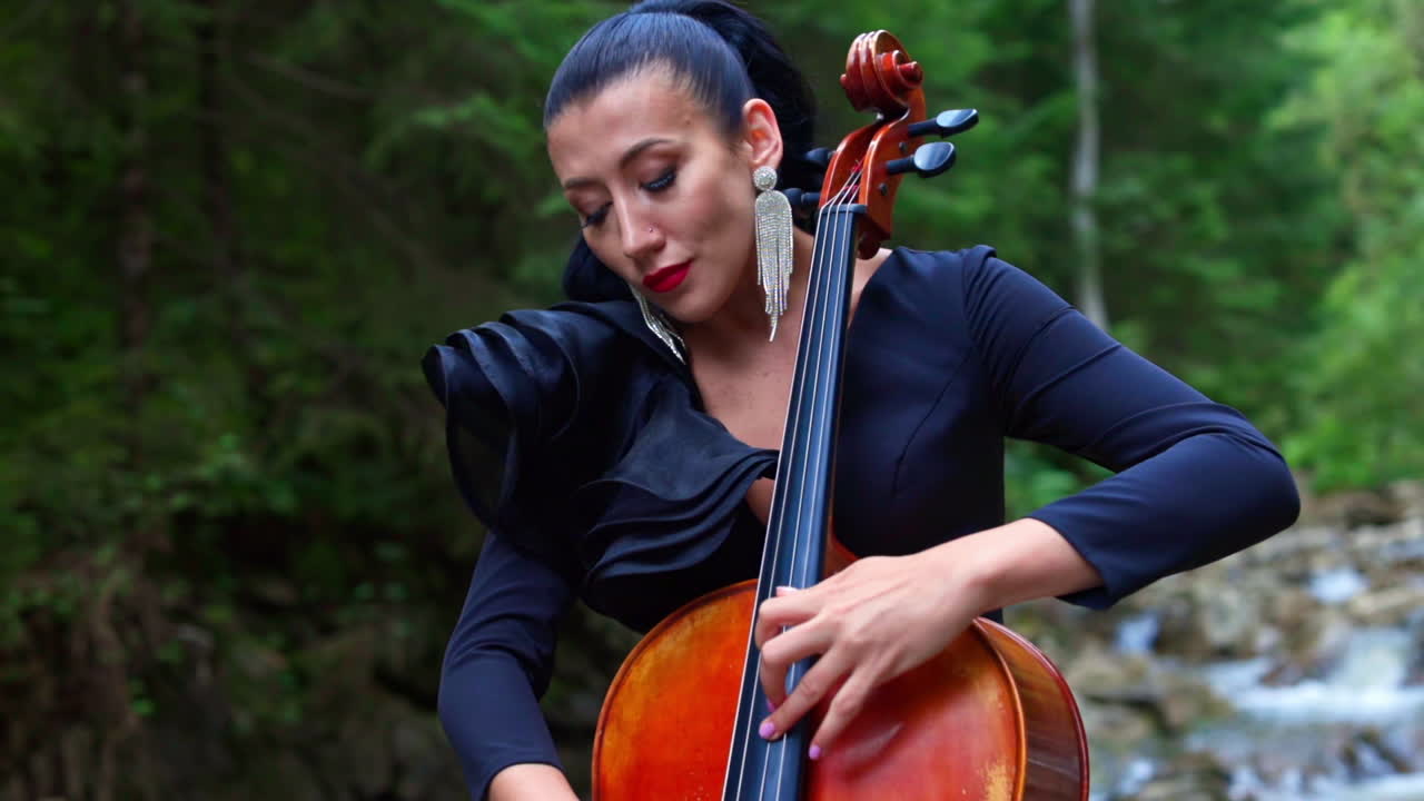 Attractive musician with cello. Beautiful woman in luxury black dress playing the musical instrument on summer nature background. Portrait of a female cellist.