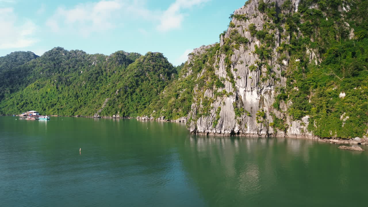 Một cảnh quay ngang mặt nước mượt mà di chuyển về phía vách đá vôi lởm chởm được bao phủ bởi cây xanh thưa thớt, với một ngôi nhà nổi màu xanh ngọc nhỏ và những dãy núi xanh tươi ở hậu cảnh dưới bầu trời quang đãng tại Hạ Long
