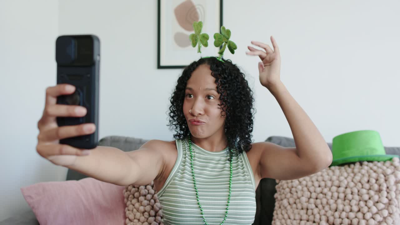 African American woman holding phone, starting call, waving, touching shamrock, laughing on sofa