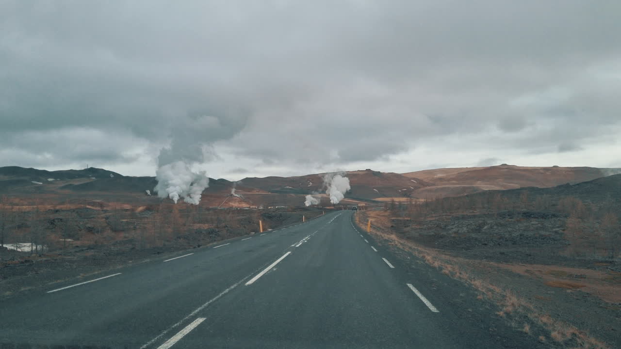 Car drives on an empty asphalt road towards a geothermal area with rising steam in Iceland