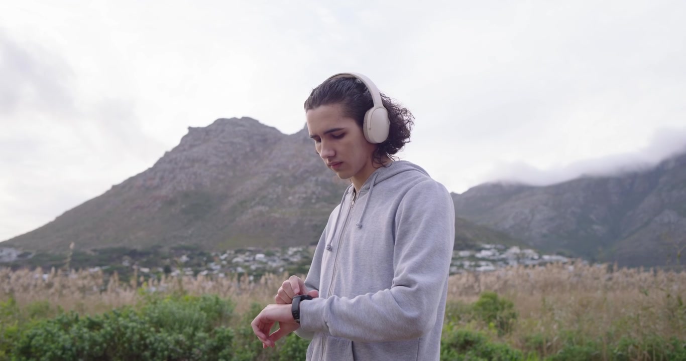 Uomo che controlla lo smartwatch, sistema le cuffie e fa jogging attraverso un campo verso una montagna per correre.