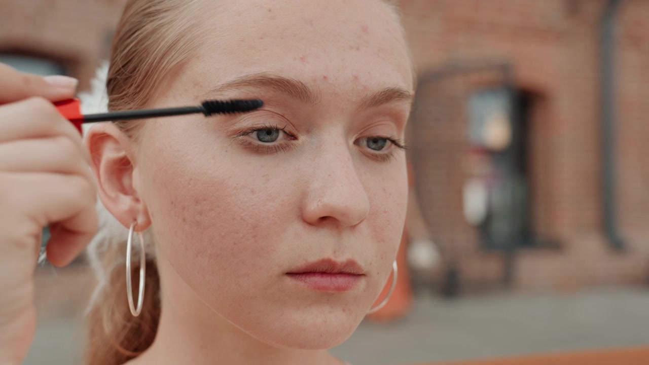 Young woman applies makeup outdoors focusing on her eye area with mascara wand. Woman wears large hoop earrings and has light colored hair tied back against urban background of brick