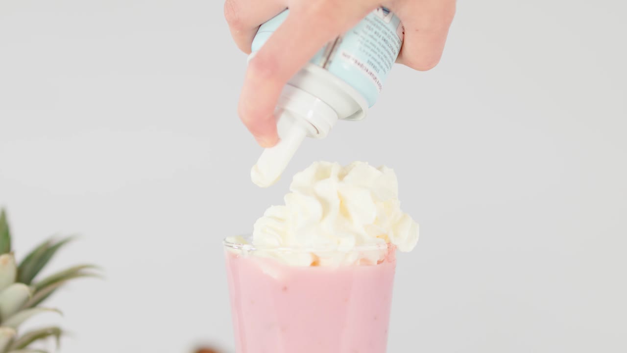 Tall pink smoothie topped with whipped cream, surrounded by fresh fruit, in bright studio lighting