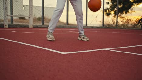 Ein-Mädchen-In-Weißen-Hosen-Lässt-Einen-Orangefarbenen-Basketball-Auf-Einem-Roten-Basketballplatz,-Der-Mit-Gittern-Umzäunt-Ist,-Frühmorgens-Bei-Sonnenaufgang-Vom-Boden-Aufprallen.