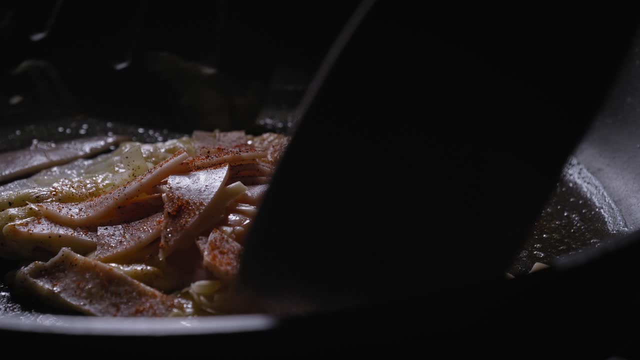 A close up shot of an egg topped with sliced ham and cheese cooking in a frying pan, the egg flipped over with a plastic spatula to ensure an even cook