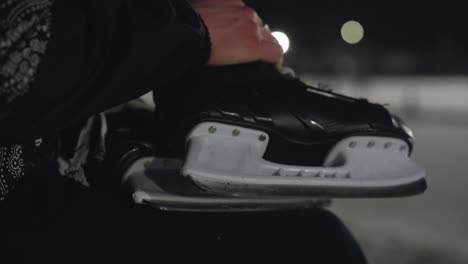 close up of person hand gripping black skating boot with detailed focus on blade and laces, set against softly blurred ice rink background under cold night atmosphere