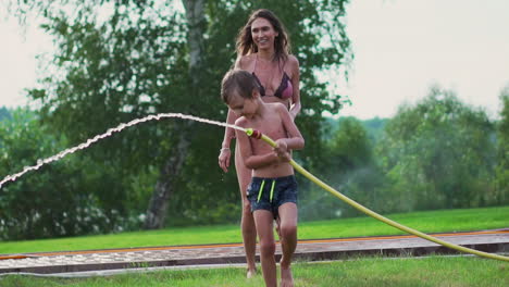 mom and son playing on the lawn pouring water laughing and having fun on the playground with a lawn on the background of his house near the lake