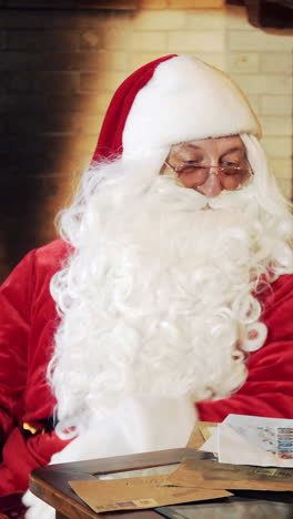 authentic santa claus with a book on fireplace backdrop. portrait of a santa in glasses reading book through a magnifying glass near the table with globe and letters at home. vertical video