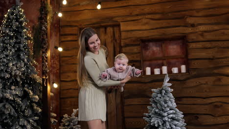 caring mom waving her cute infant baby. mother playing with her son in the wooden hut. christmas trees at backdrop.
