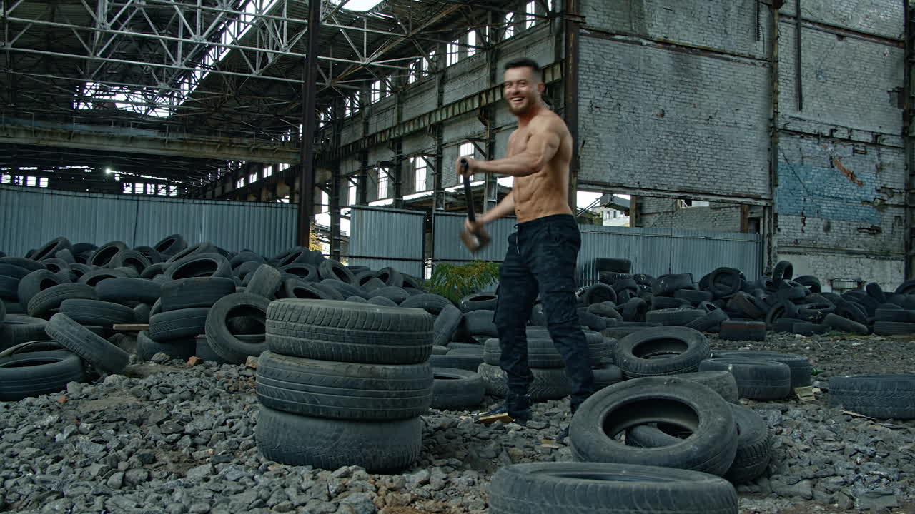 Sportsman with a heavy hammer in old plant. Sporty man trying to break pile of car tires with a metal hammer and smiles to camera. Workout in abandoned place.