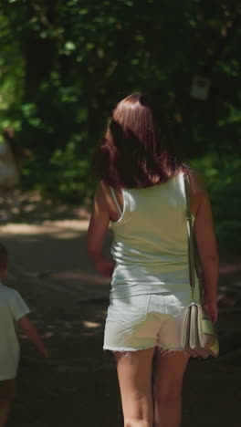 young woman with toddler boy walks along shady road across forest slow motion. mother with little son enjoys weekend in nature park. family vacation