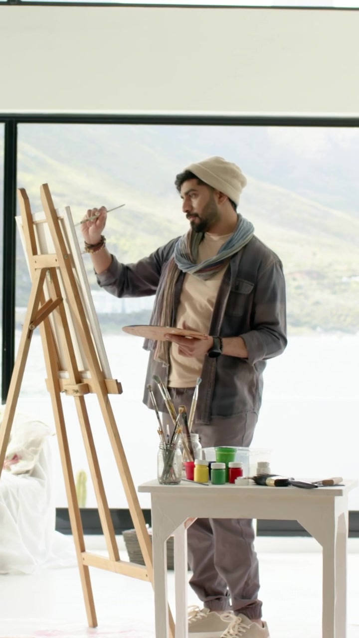 Vertikal video: Malende kunstner med hat kigger på palet, dypper pensel i farve ved staffeli i studie.