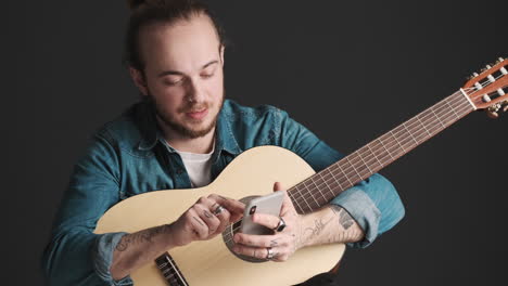 caucasian young man scrolling on smartphone while holding guitar.