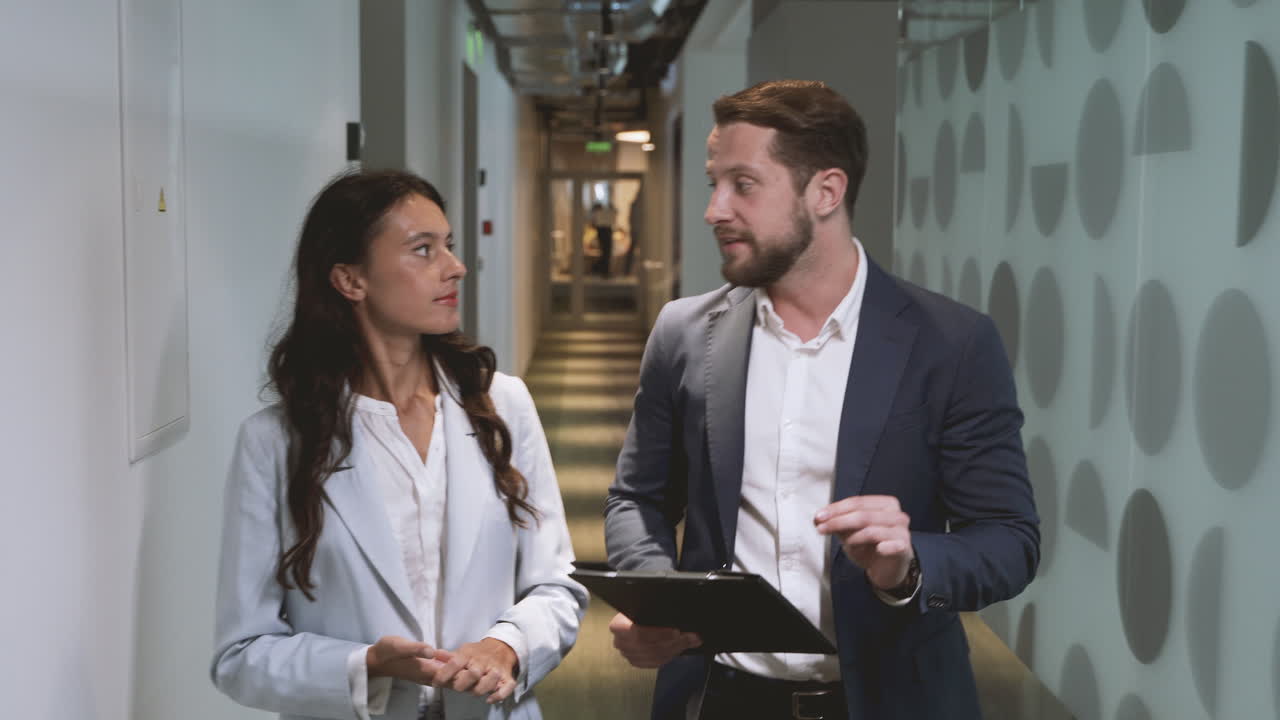 A young businessman and a young businesswoman walk through the corridors of an office building discussing company matters.