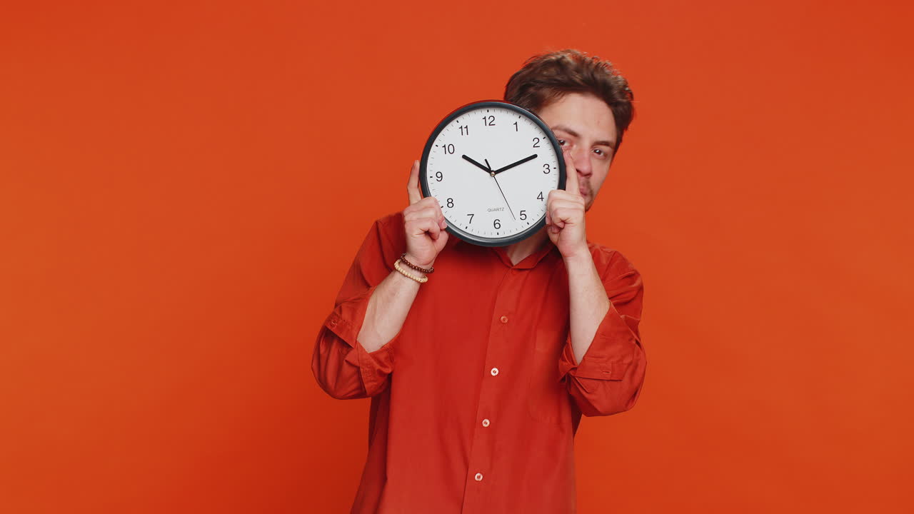 Serious strict young man holding office clock hiding behind checking time on watch obscuring face