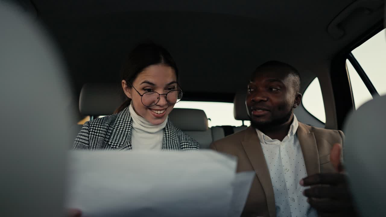 Happy brunette girl in round glasses communicates with her colleague a man with Black skin in a brown jacket while they review their plans and thoughts on paper while traveling in a modern salon in a car outside the city