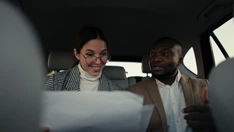 Happy-brunette-girl-in-round-glasses-communicates-with-her-colleague-a-man-with-Black-skin-in-a-brown-jacket-while-they-review-their-plans-and-thoughts-on-paper-while-traveling-in-a-modern-salon-in-a-car-outside-the-city