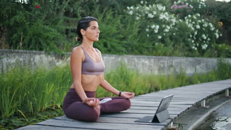 señora tranquila sentada en posición de loto banco del parque. mujer viendo lección de yoga en línea